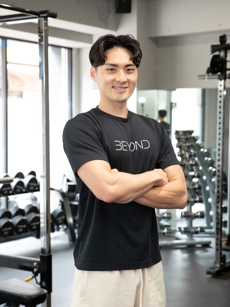 BEYOND武蔵小杉店のパーソナルトレーナーがジム内で腕を組んで笑顔で立っている
