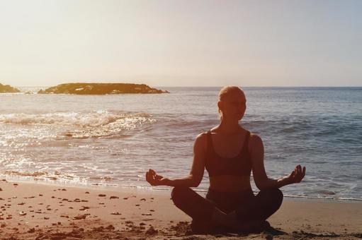 海辺で朝日を浴びながら瞑想する女性。リラックスと深呼吸によってミトコンドリアを活性化するイメージ。