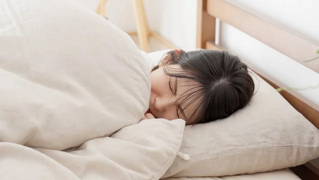 「布団にくるまって眠る女性。冬の冷えや寝起きの悪さをイメージした写真」