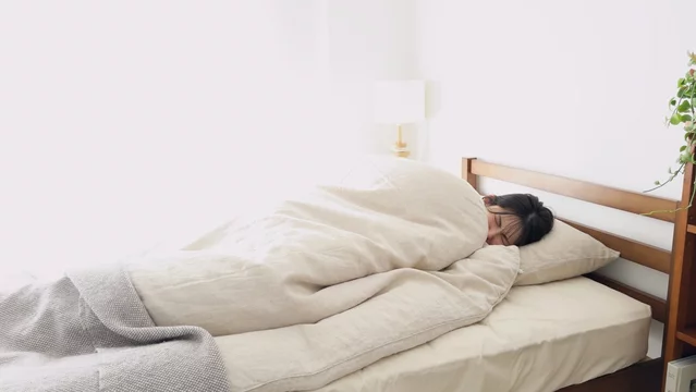 「冬の朝に寒さで布団から出られず縮こまっている女性」